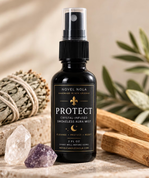 A 2 fl oz black bottle of Novel Nola Protect Crystal-Infused Smokeless Aura Mist, styled on a stone surface with crystals, a sage bundle, and wooden tools against a warm beige background.